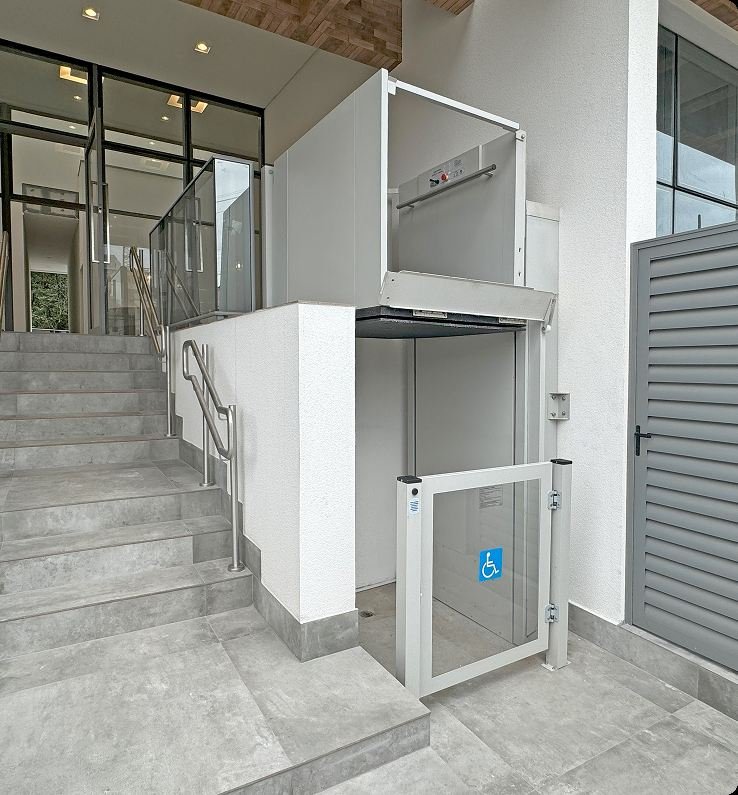 Plataforma vertical de acessibilidade instalada ao lado de uma escada, com portão de vidro.